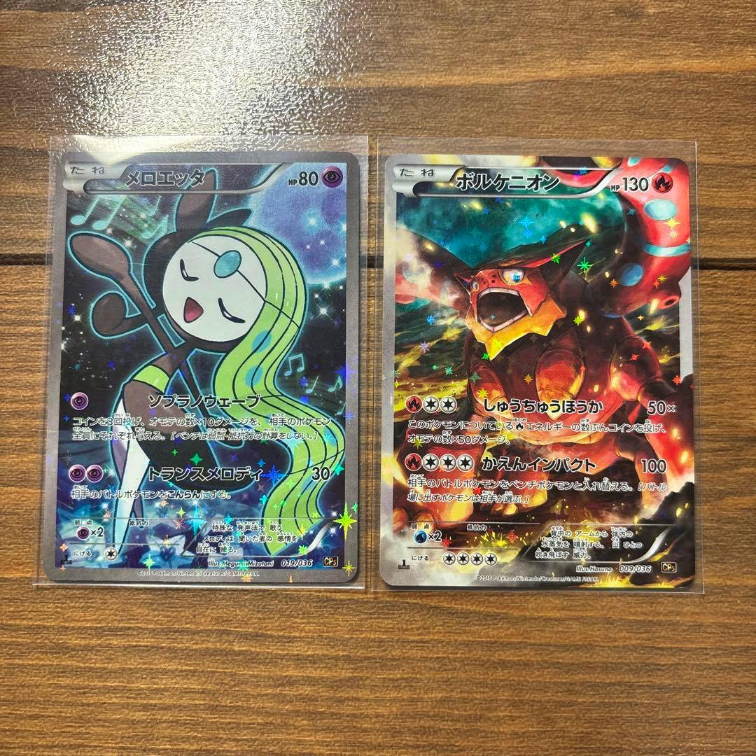 Two sleeved Pokémon trading cards side-by-side on a wooden surface showing the front art of メロエッタ (left) and ボルケニオン (right).