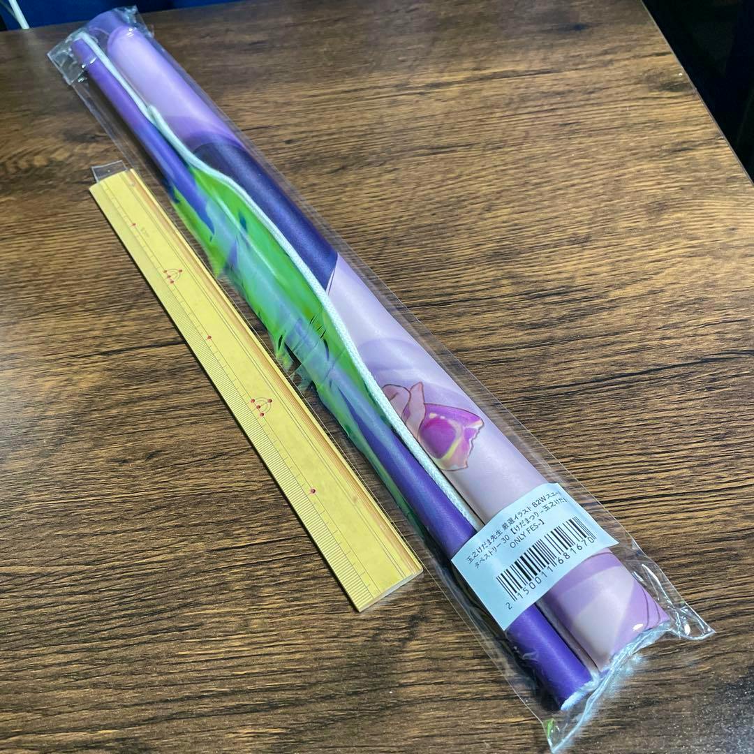 A rolled B2 tapestry in clear plastic packaging shown on a wooden surface next to a yellow ruler.