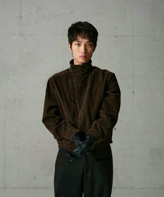 Male model wearing a brown button faux nubuck blouson jacket against a gray concrete wall.