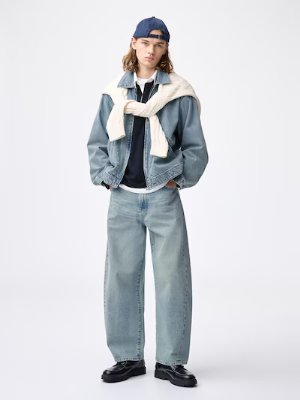 Male model wearing light-wash barrel leg jeans styled with a jacket and cap against a white background.