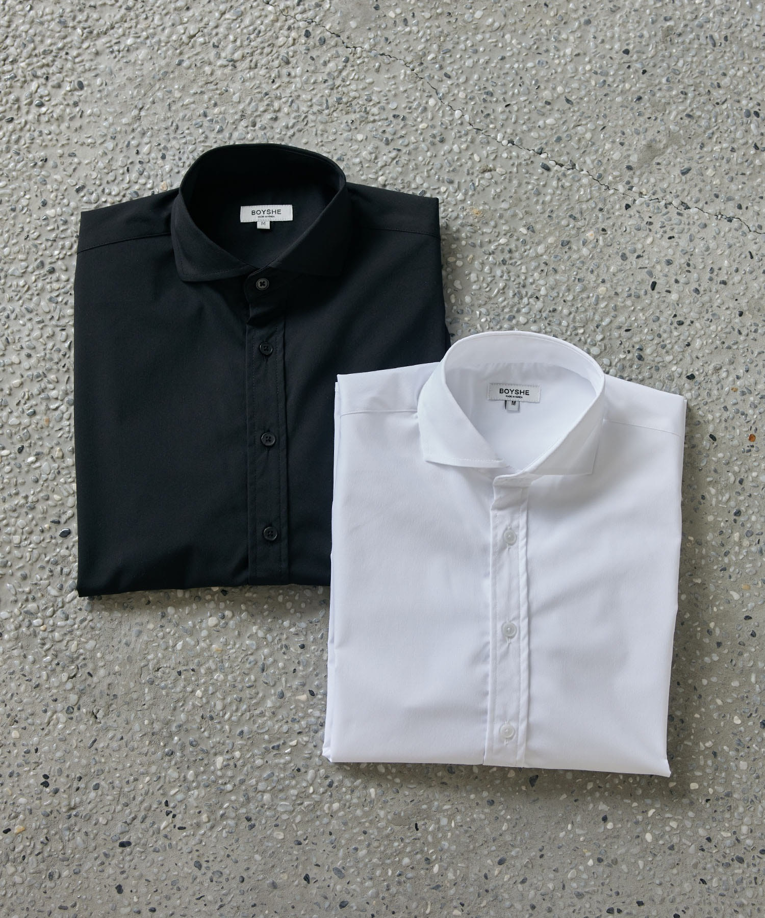 Two folded men's cutaway-collar dress shirts, one black and one white, photographed on a speckled concrete surface.