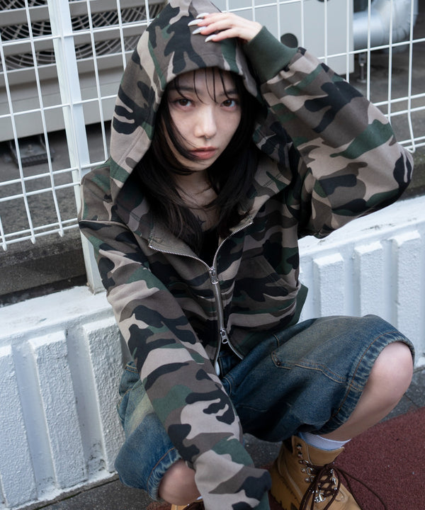 Model wearing a camouflage short zip hoodie with the hood up, seated against a white metal fence.