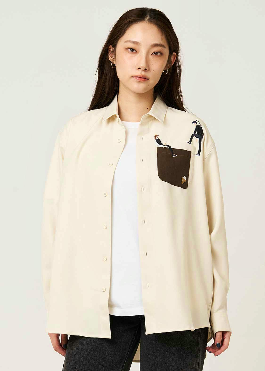 A men's cream loose-fit button-up shirt worn by a model, showing a dark chest pocket with small illustrated figures.