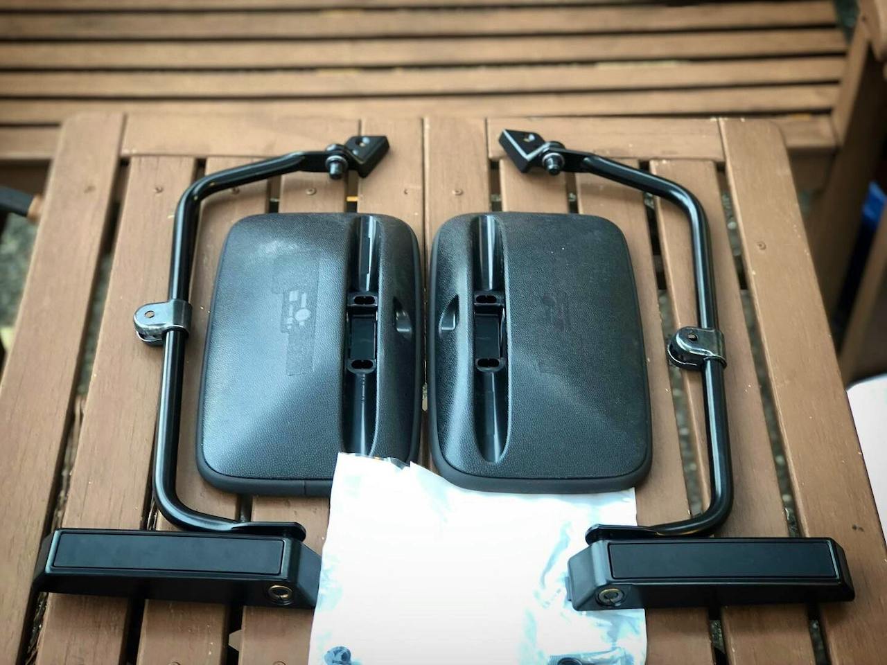 Two black car side mirrors with mounting arms laid side by side on a wooden surface, matching a Mercedes G-Class W463 style.