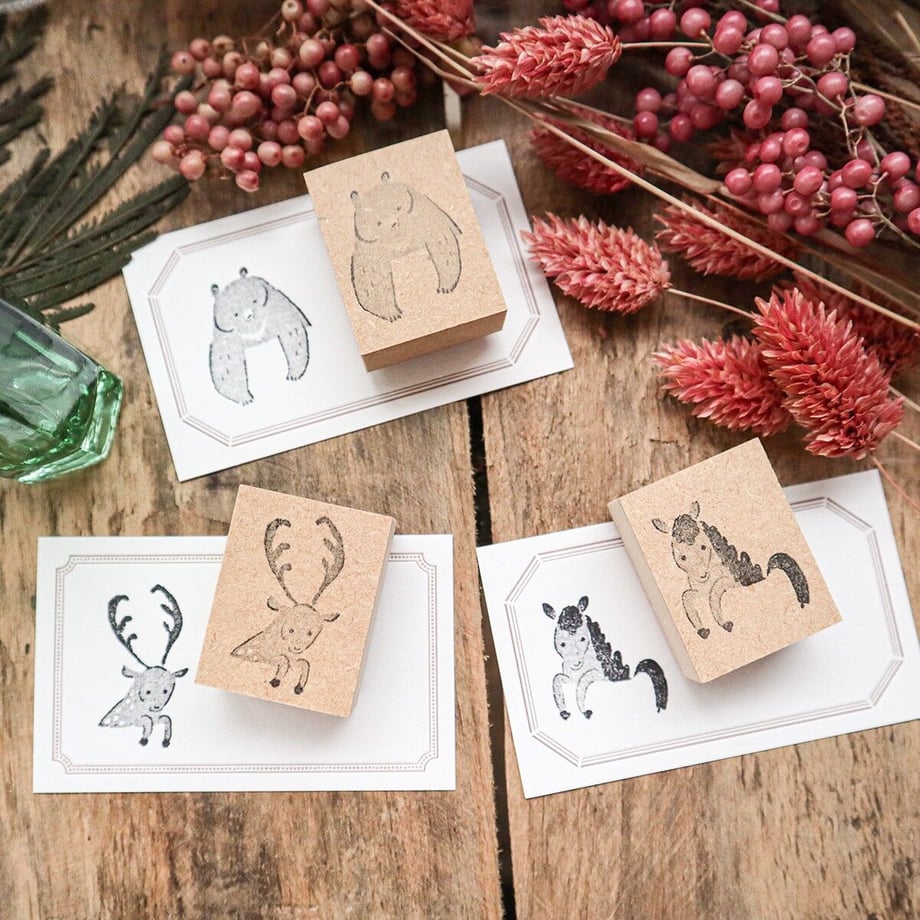 Four wooden stamp blocks showing animal designs alongside their stamped impressions on cards, arranged on a wooden surface with dried flowers.