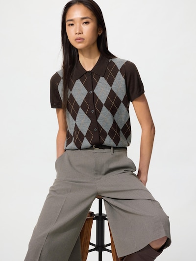 Model wearing the merino-blend polo cardigan with an argyle front, seated on a stool against a white background.