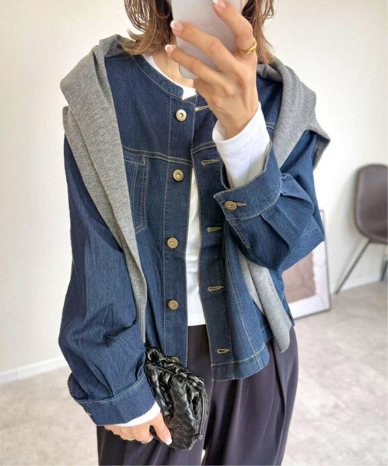 Model wearing a blue denim blouson jacket with visible buttons and cuffed sleeves, layered with a gray sweater draped over the shoulders and holding a black woven clutch.