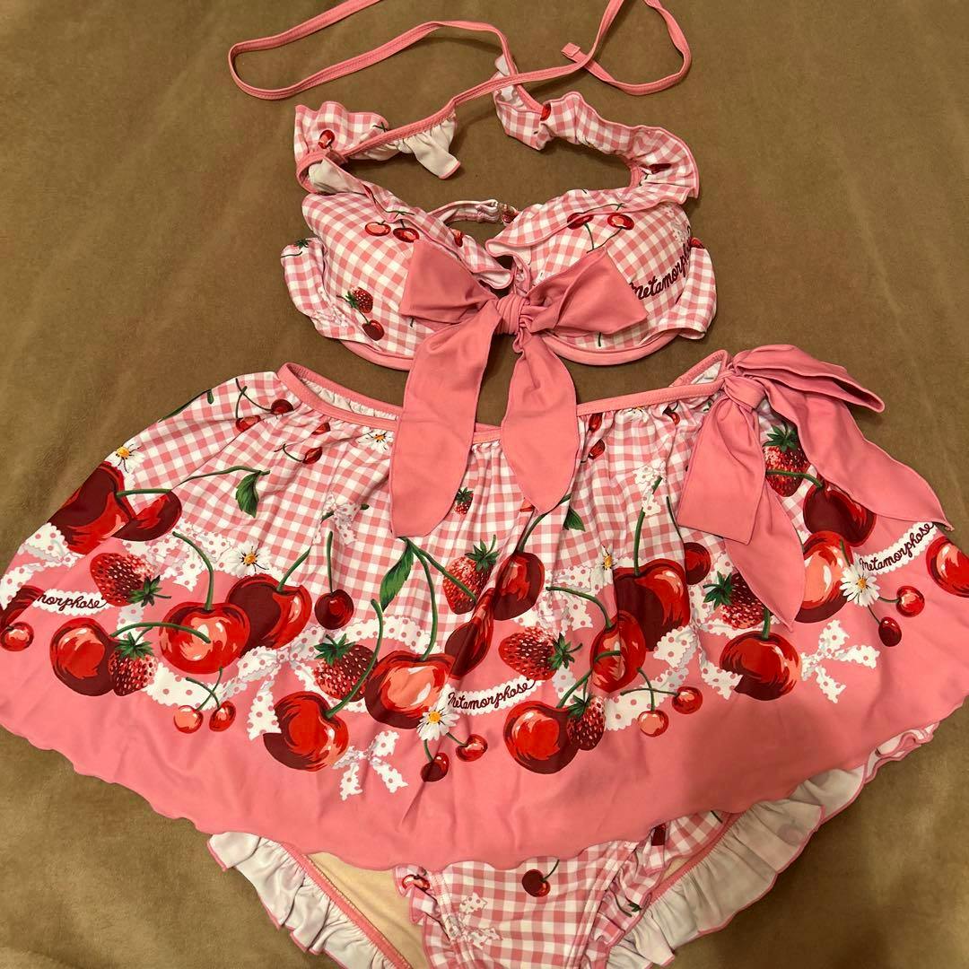 Pink Metamorphose two-piece women's swimsuit with cherry print, bow, and skirt laid flat on a brown surface.