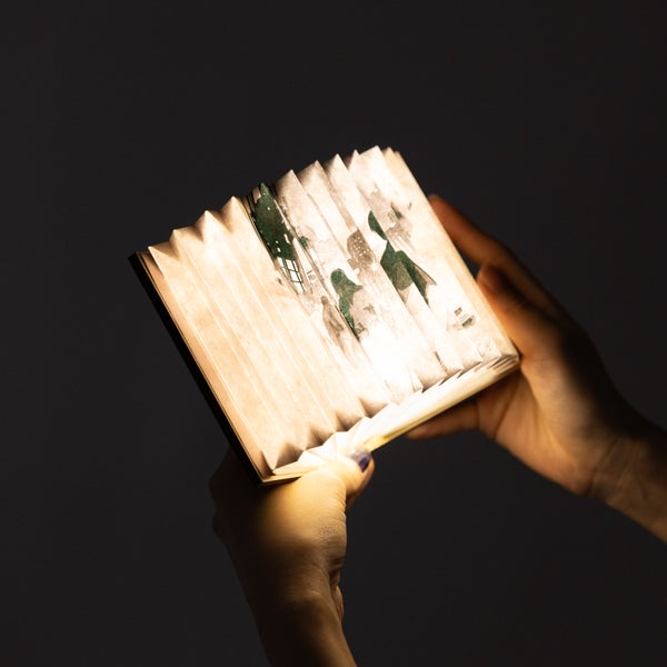 Hands holding an open, illuminated book-shaped desk lamp against a dark background.