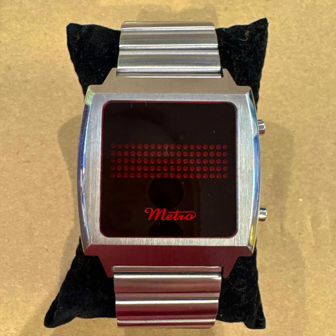 Front view of the Metro LED digital wristwatch showing the red dot-matrix display, Metro logo, and stainless steel case and bracelet on a black cushion.
