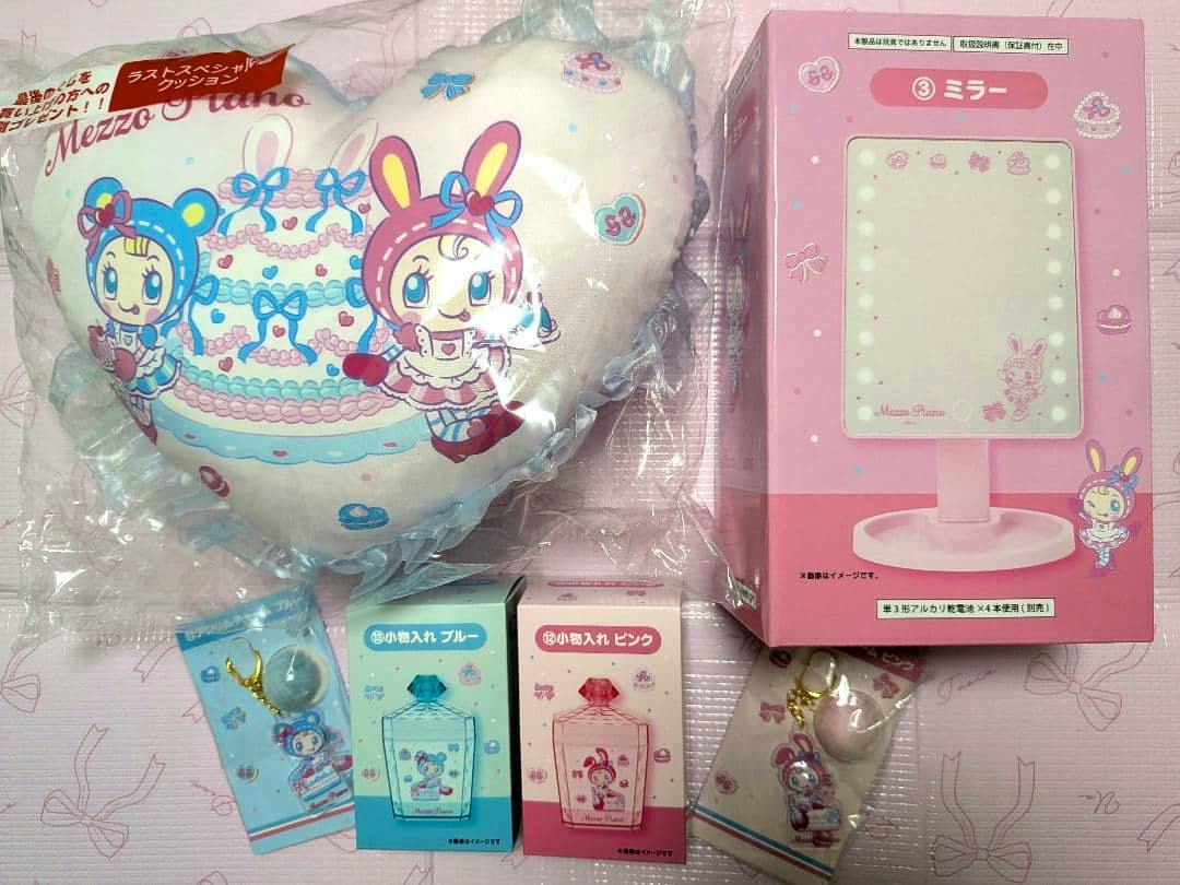 A photo of a Mezzo Piano Ichiban Kuji 6-piece goods set showing a heart-shaped cushion in plastic, a boxed pink mirror, two small accessory boxes, and two packaged keychains.