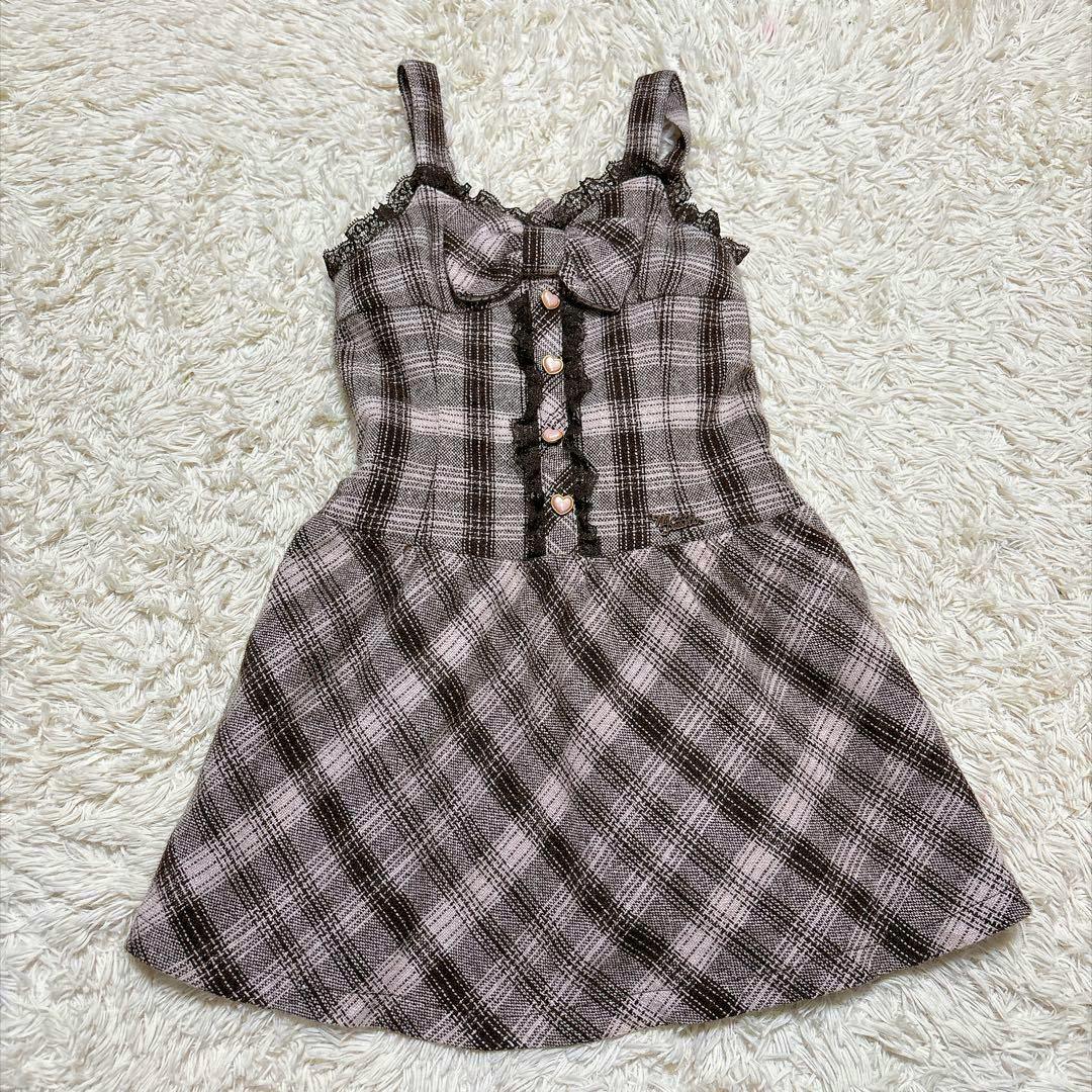 Pink and brown check patterned jumper skirt with straps, bow, heart buttons, and lace trim laid flat on a white rug.