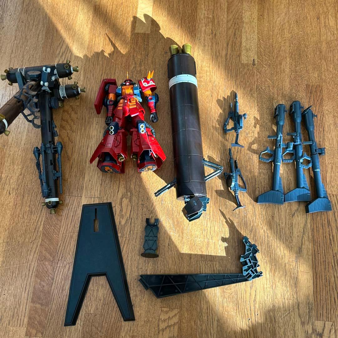 Disassembled MG 1/100 ガンプラ ジャンク サイコザク ver.ka model parts and accessories laid out on a wooden floor, including a red Psycho Zaku torso and multiple weapons and stands.