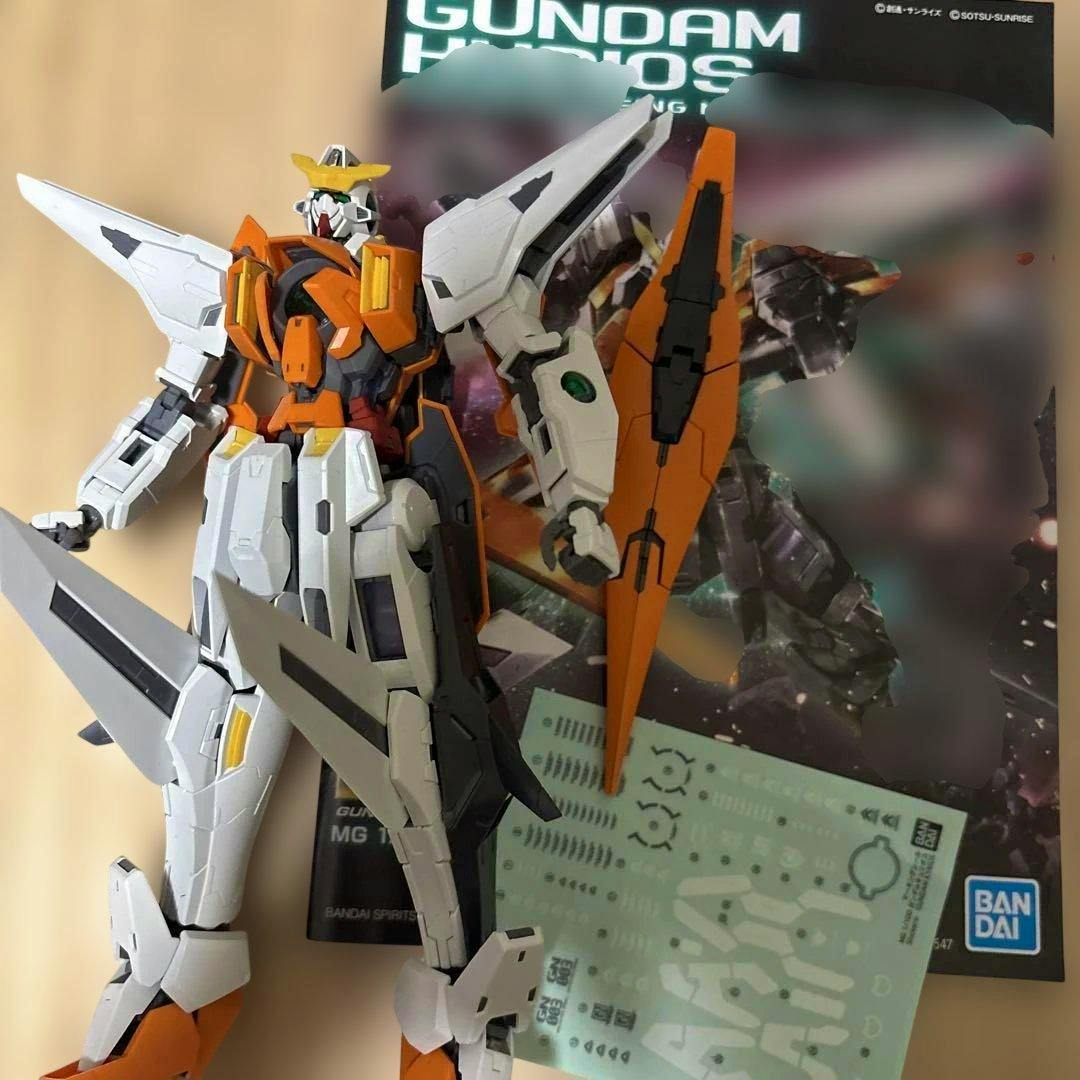Assembled MG Gundam Kyrios model (completed kit) posed in front of its box with a decal sheet visible.