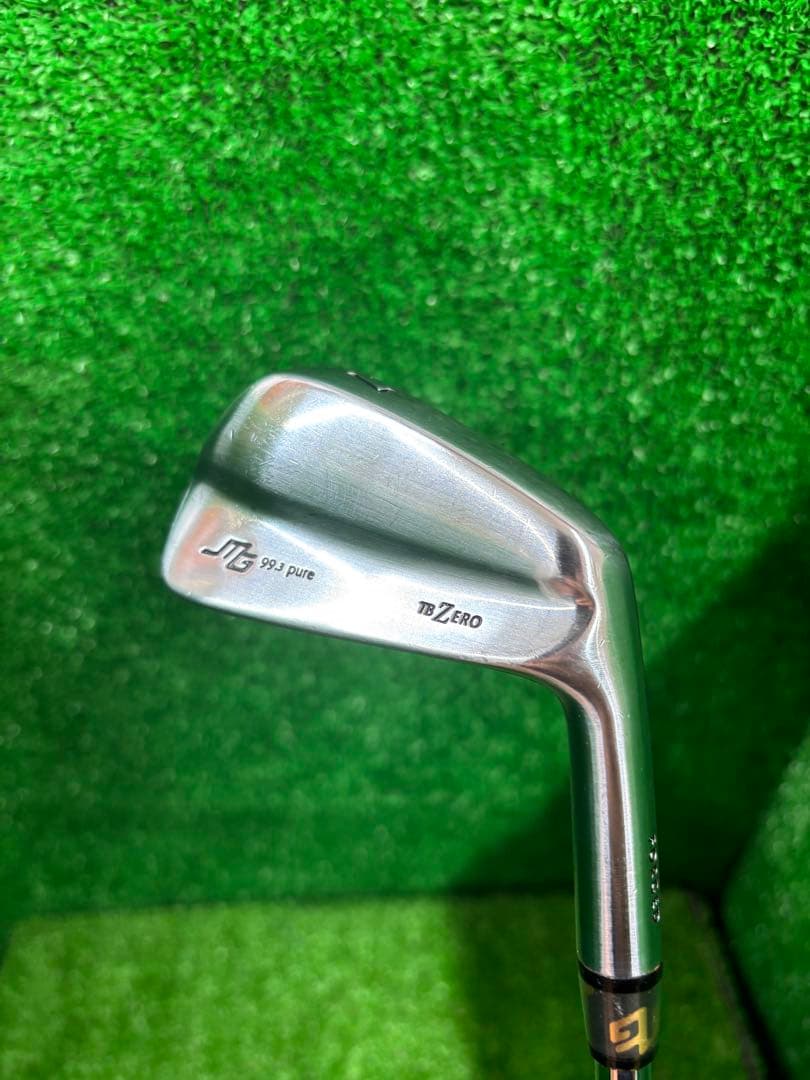 Close-up photo of a TB ZERO iron club head showing the 'TB ZERO' marking against a green turf background.