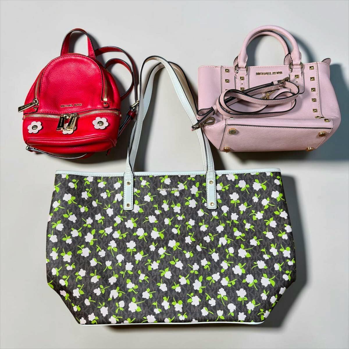 Three Michael Kors bags — a floral-print tote, a pink studded handbag, and a small red backpack — laid flat on a light background.