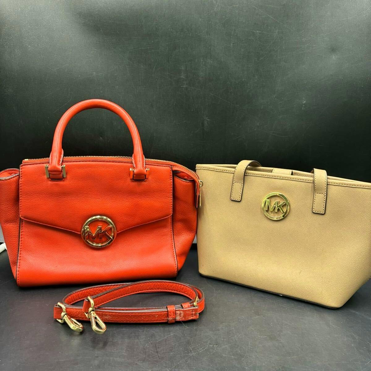 Two Michael Kors bags: an orange-red top-handle bag with a detachable strap and a beige tote with an MK logo, shown on a dark surface.