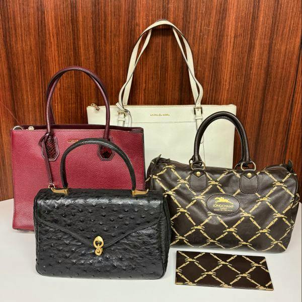 Four handbags arranged together: a cream Michael Kors tote, a burgundy bag, a black ostrich-textured top-handle bag, and a brown Longchamp-patterned bag.