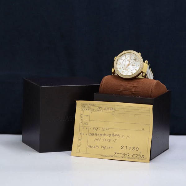 Gold Michael Kors wristwatch displayed on a brown cushion with its box and a yellow paper next to it.