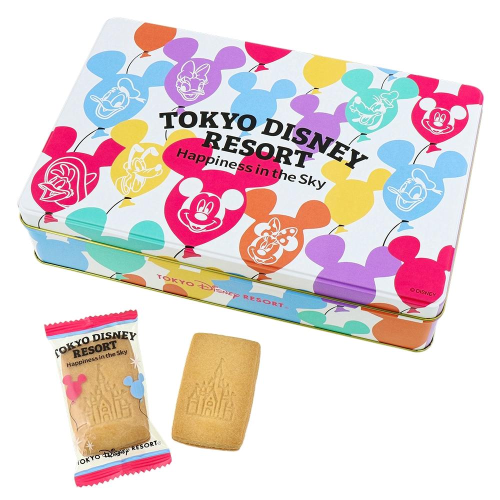 A colorful Tokyo Disney Resort cookie tin decorated with Mickey-shaped balloon silhouettes, shown with two cookies (one wrapped).