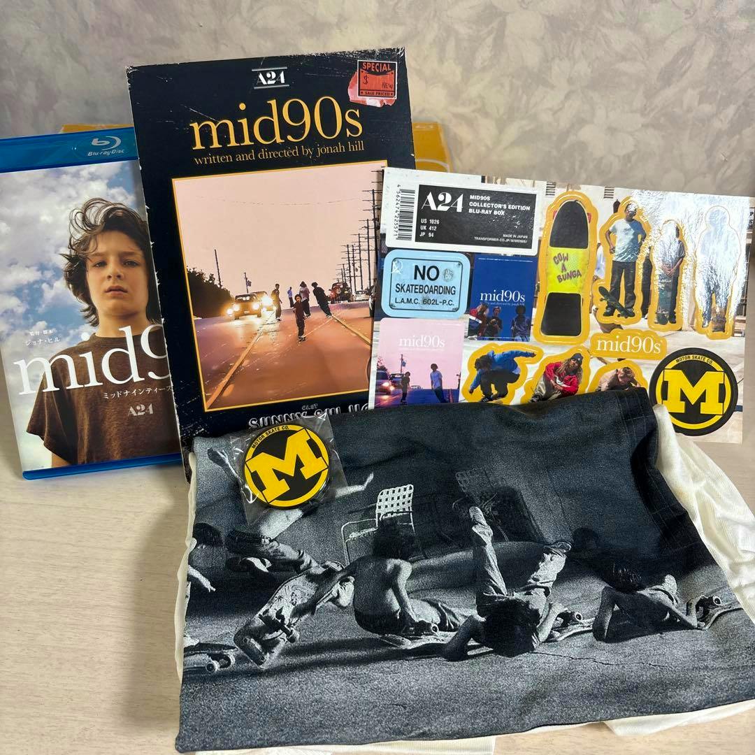 Photo of the mid90s set showing the Blu-ray case and box, sticker sheet, round yellow "M" badge, pamphlet, and a folded white T-shirt with a black-and-white photo print.