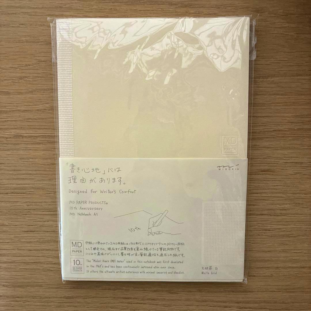 A wrapped MD Notebook A5 10th-anniversary edition with cream cover and a paper band showing writing samples and a grid.