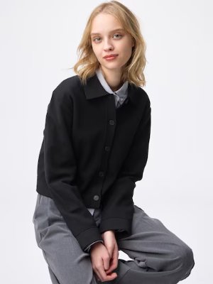 A model seated wearing a black polo-style cardigan buttoned over a collared shirt against a white background.