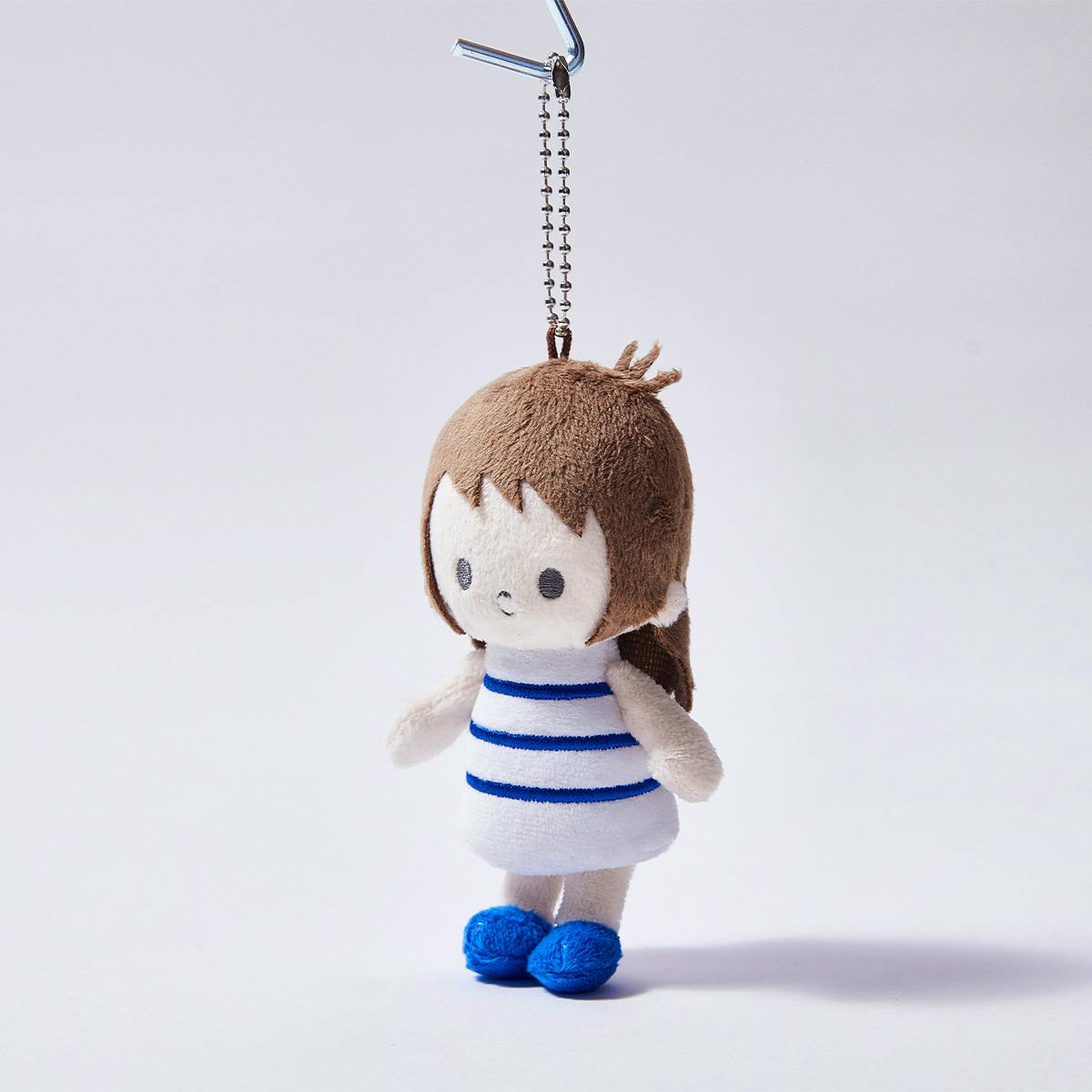 A plush keychain of a small girl character with brown hair wearing a white dress with blue stripes and blue shoes, hanging from a metal ball chain.