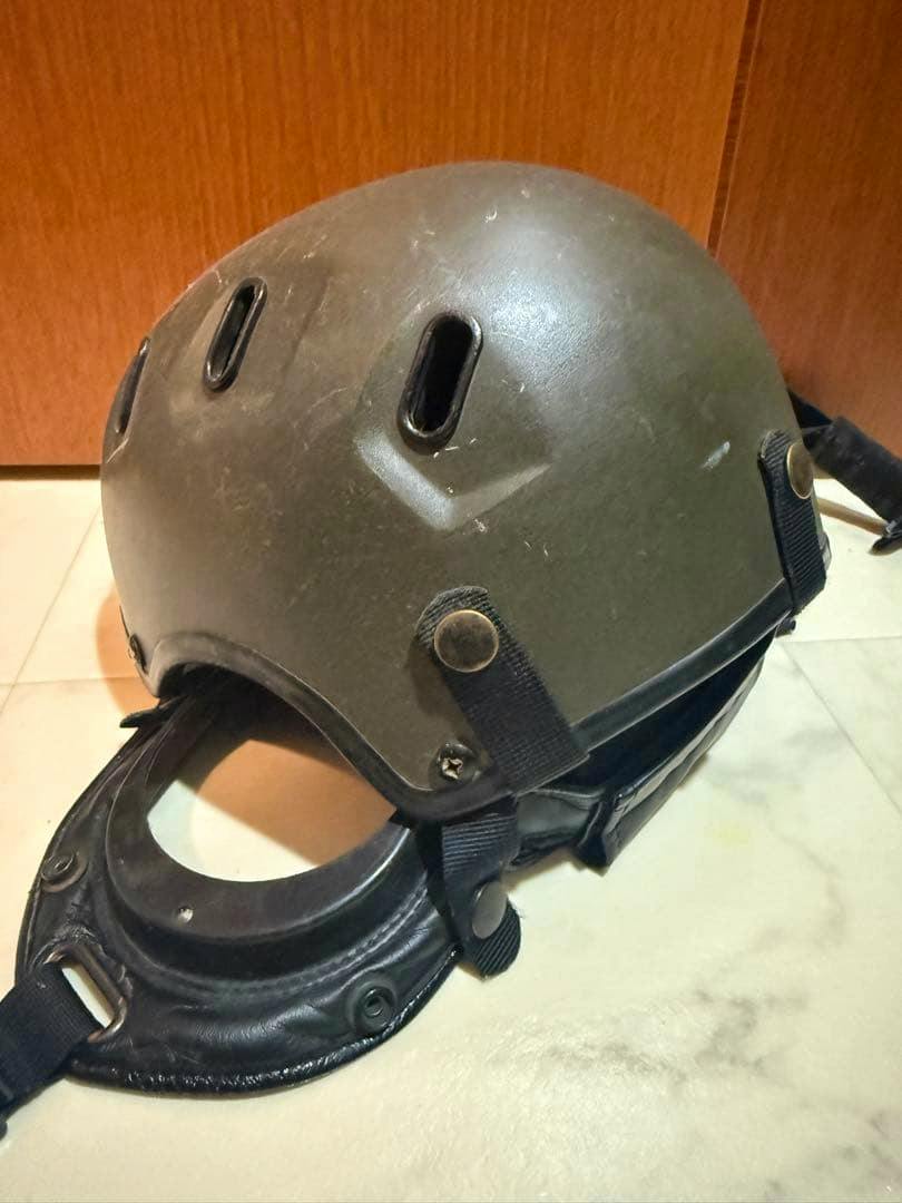 Green military helmet with attached chin strap and leather neck guard sitting on a tiled surface in front of a wooden background.
