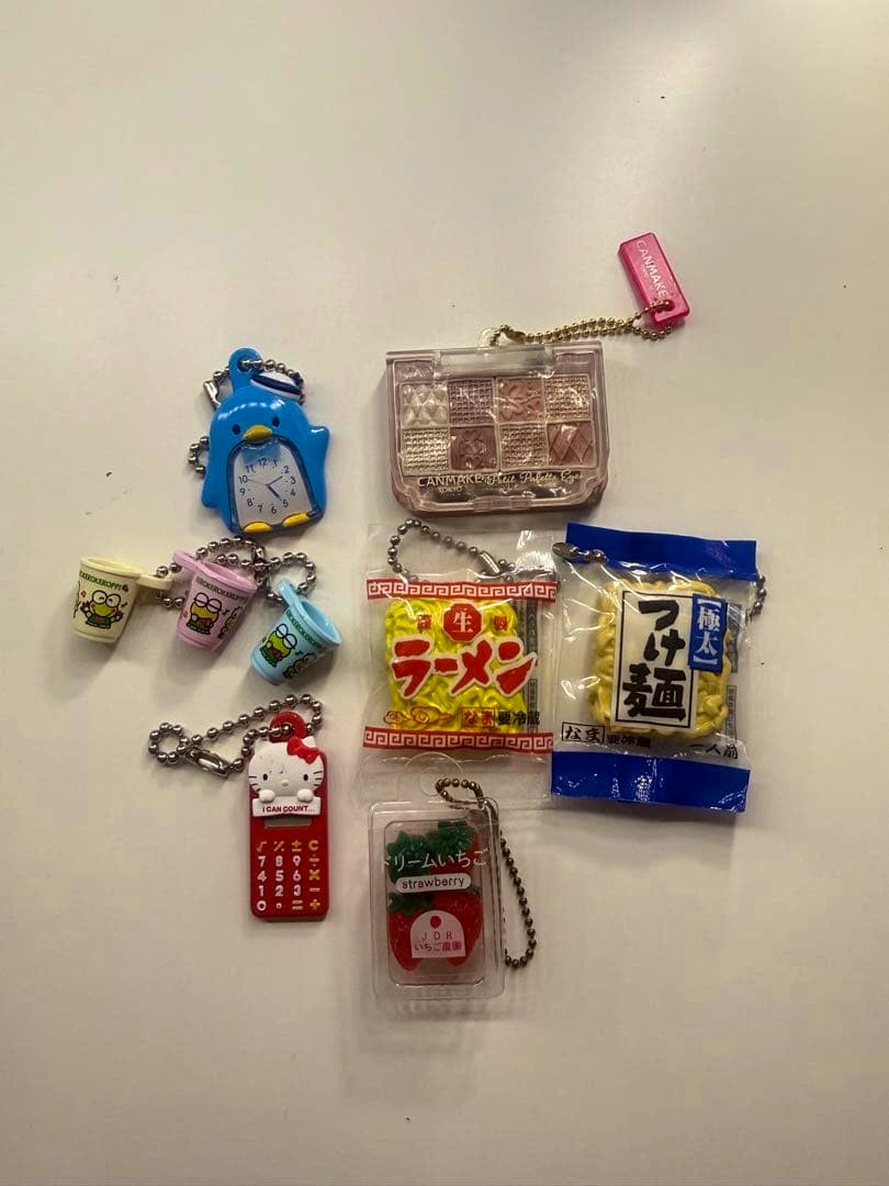 Assorted miniature gacha keychain toys laid on a light surface, including a penguin clock, small ramen and noodle packets, a tiny makeup palette, mini cups, a Hello Kitty phone, and a strawberry container.