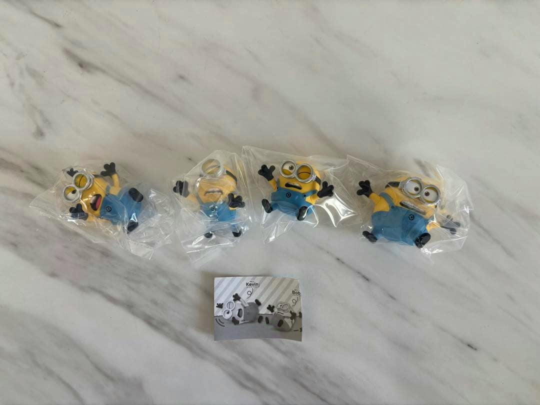 Four Minions plastic figures in individual clear bags displayed on a marble surface with a small paper insert.