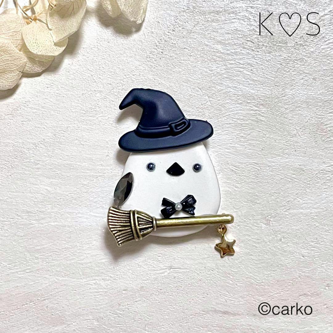 A waist buckle depicting a white bird wearing a black witch hat and holding a broom with a small star charm on a textured white background.