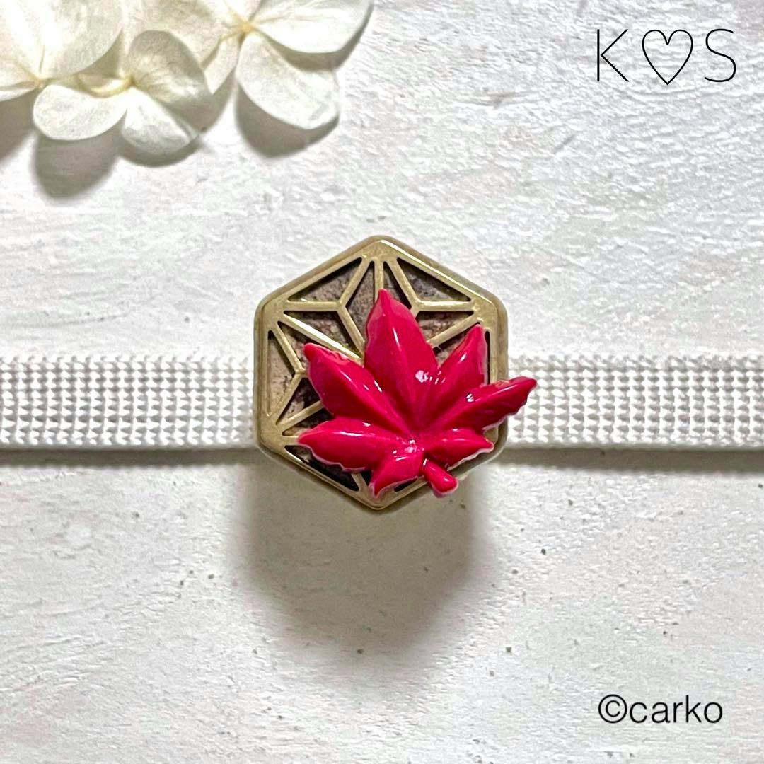 Hexagonal alloy waist buckle with a red maple-leaf ceramic ornament threaded on a white cord against a textured background.