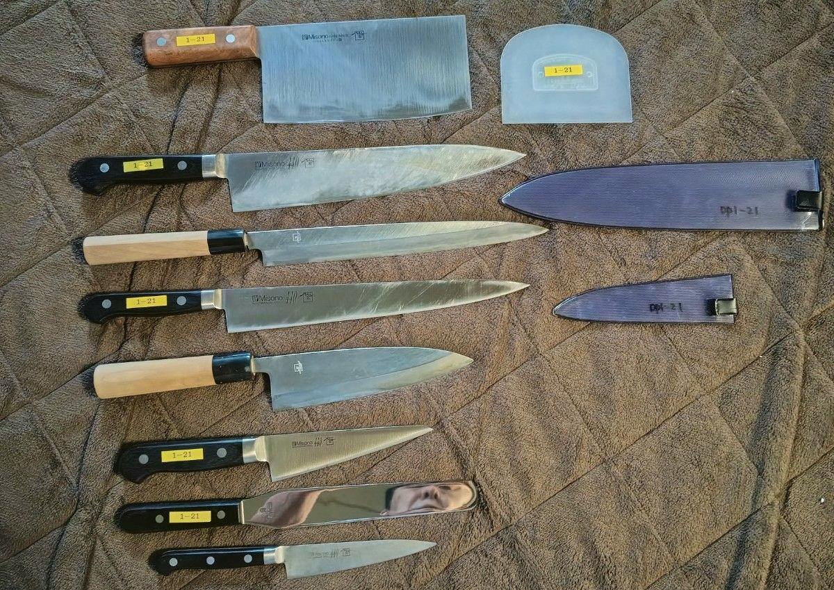 Seven Misono chef knives with various handles and two sheaths laid out on a brown quilted surface.