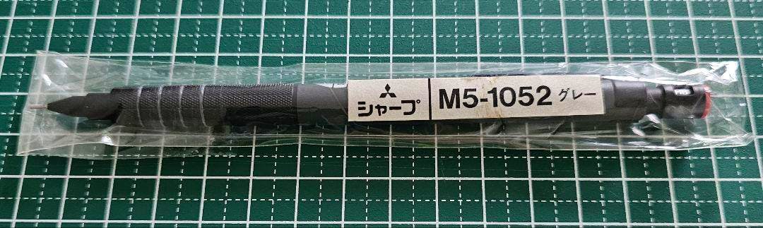 Sealed 三菱 uni シャープペンシル M5-1052 mechanical pencil in plastic wrap on a green grid cutting mat.