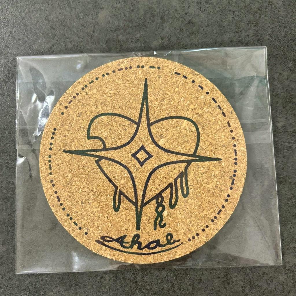 A round cork coaster with a stylized three-pointed star design and dotted border, shown in clear plastic packaging on a dark surface.