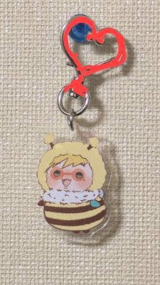 Acrylic keychain featuring a small character in a bee-striped outfit attached to a red heart-shaped connector against a textured wall.