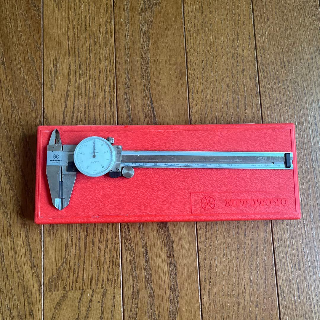 A Mitutoyo dial caliper with a white dial placed on a red Mitutoyo case on a wooden floor.