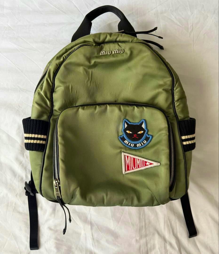 Front view of a khaki Miu Miu backpack showing a cat patch and a triangular MIU MIU pennant on the front pocket.