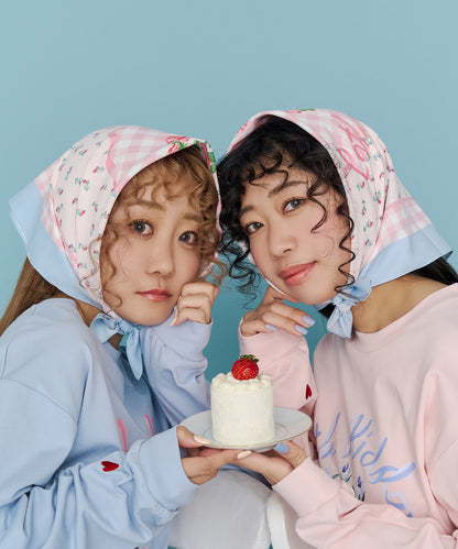 Two women wearing pink patterned scarves tied under their chins hold a small cake on a plate against a blue background.