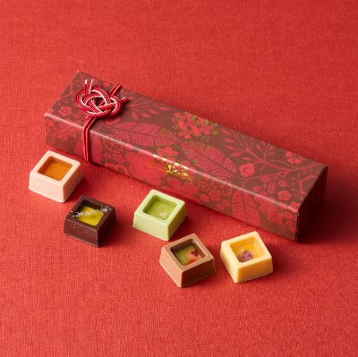 A decorative red gift box with five square fruit-and-honey confections arranged on a red surface.