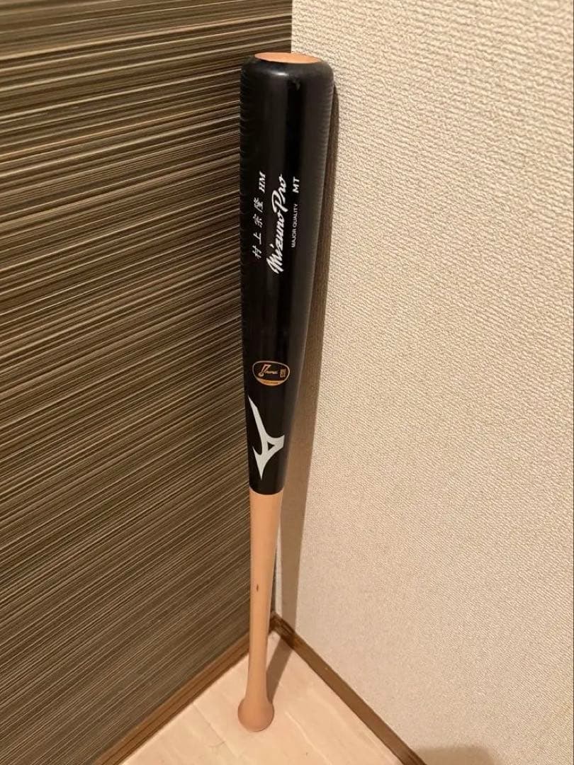 A Mizuno Pro baseball bat with a black barrel and natural wood handle standing upright in a room corner.
