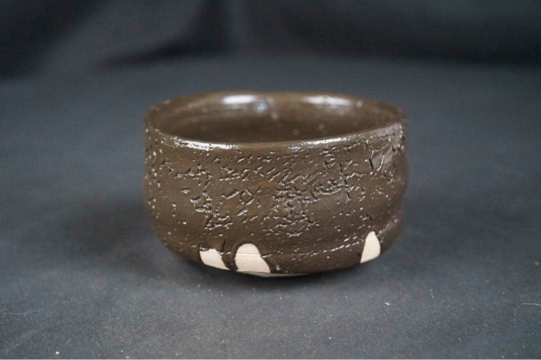 Black-glazed Japanese tea bowl (chawan) with textured surface and exposed clay spots on a dark background.