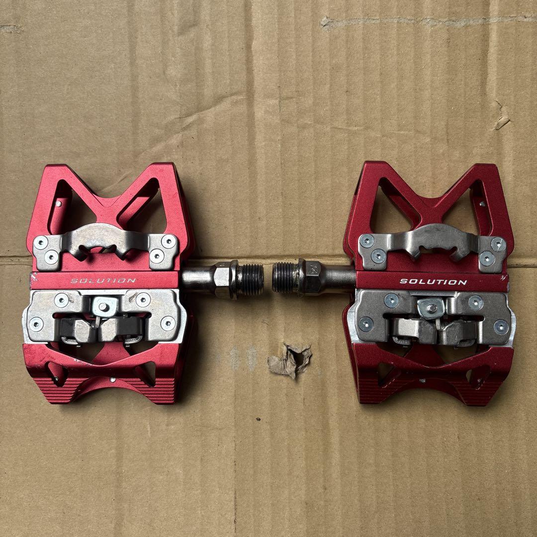 Pair of red MKS Solution bicycle binding pedals laid on cardboard showing the clip mechanisms and spindle.