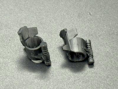 Two small dark plastic figure greave parts on a gray surface.