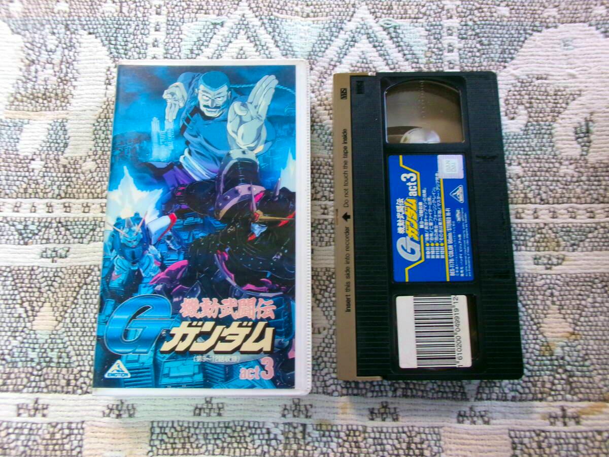 A VHS cassette and its illustrated case for the Japanese anime "機動武闘伝 Gガンダム act.3" displayed side by side on a patterned cloth.