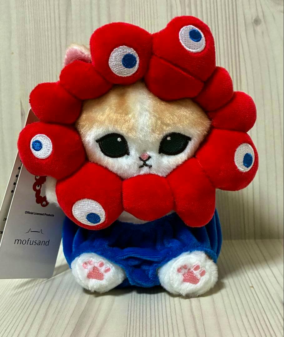 Front view of the mofusand EXPO2025 plush mascot with a red petal-like headpiece, blue shorts, and a visible tag.