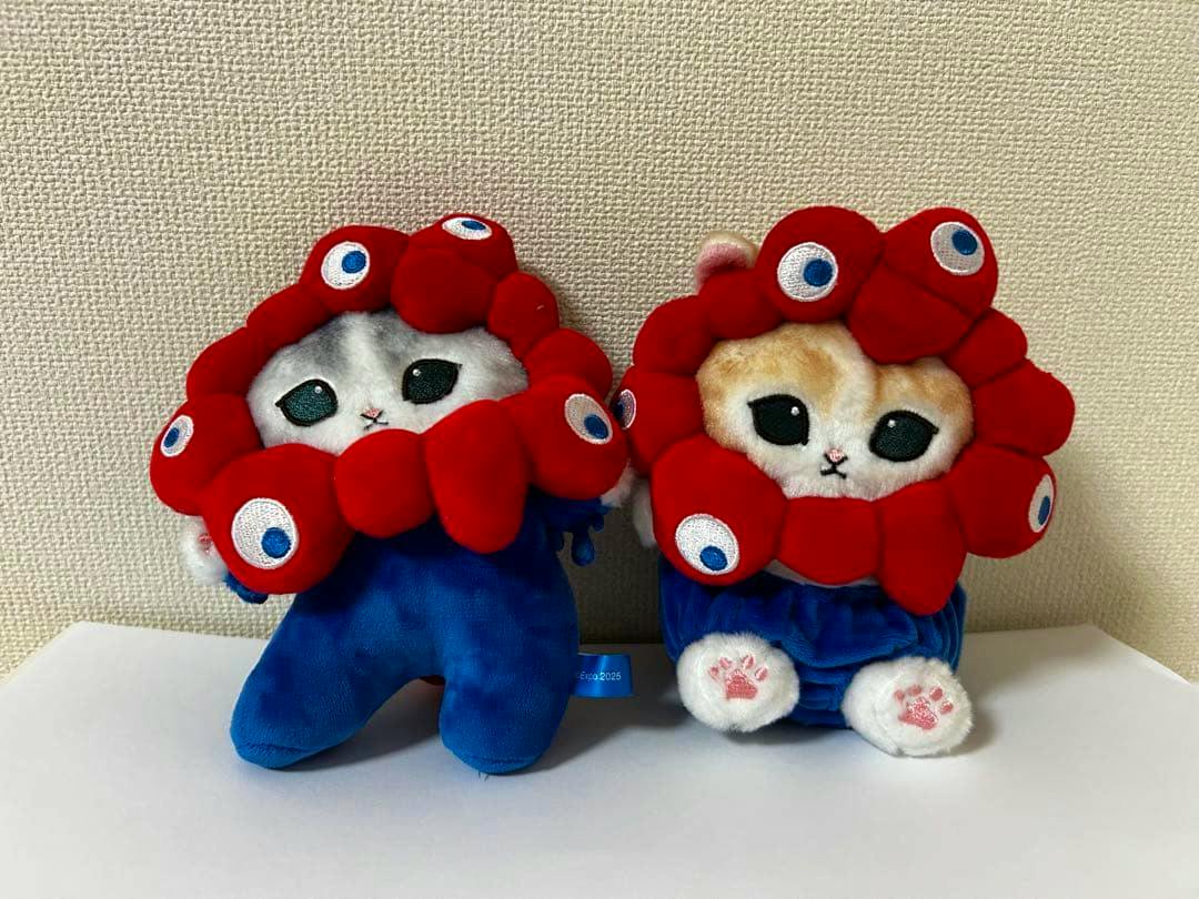 Front view of two mofusand plush toys wearing red flower-like hoods and blue bodies seated on a white surface.