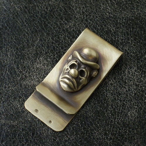 A metal money clip featuring a sculpted pierrot clown face placed on a textured dark surface.