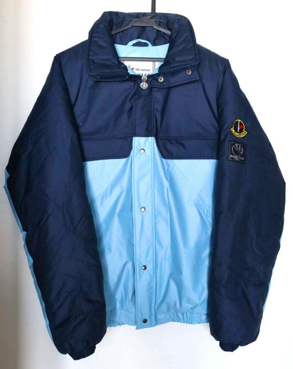 Front view of the men's nylon Moncler jacket in navy and light blue with sleeve patches, hanging on a hanger.